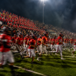 A football team in red charges onto the field beneath stadium lights as a red-uniformed marching band and red-and-black hometown crowd cheer from the stands.