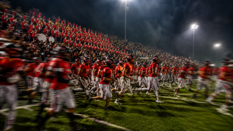 A football team in red charges onto the field beneath stadium lights as a red-uniformed marching band and red-and-black hometown crowd cheer from the stands.